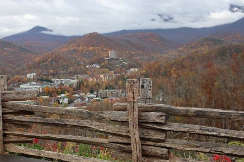 A wooden fence in the foreground overlooks a valley filled with autumn-colored trees and a small town below.