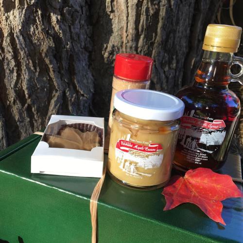 A collection of maple products including syrup, cream, and candies, displayed against a tree backdrop with a fall leaf.