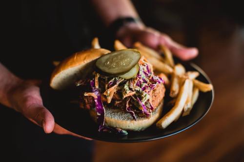 A person holds a plate with a fried chicken sandwich topped with coleslaw and pickles, alongside crispy fries.