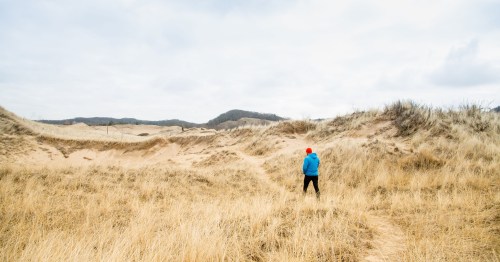 A person in a blue jacket and red hat walks through sandy dunes with tall grass under a cloudy sky.