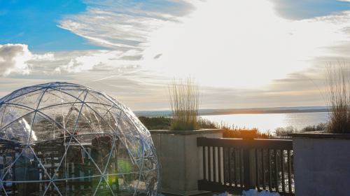 A clear geodesic dome on a terrace overlooking a shimmering lake and a bright sky with clouds.