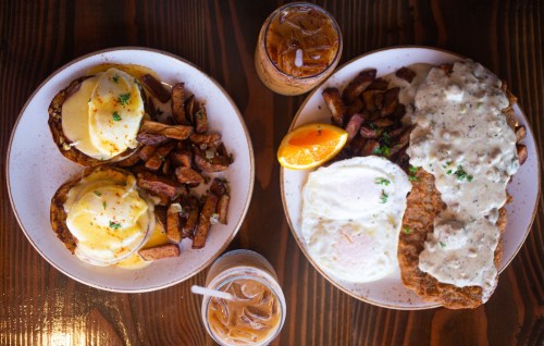 A table with two plates of breakfast: eggs Benedict with potatoes and a plate of fried chicken, eggs, and gravy.