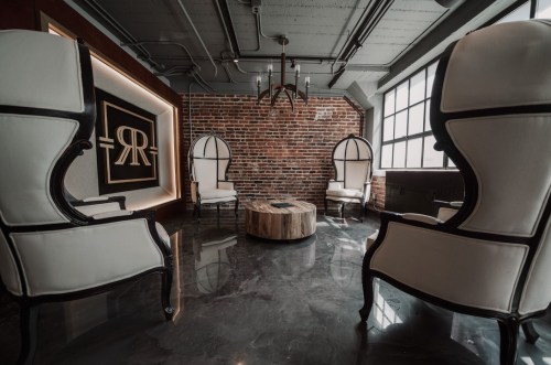 A stylish lounge with two elegant chairs, a wooden table, and a brick wall featuring a logo. Modern and inviting atmosphere.