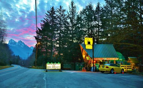 A roadside diner with a yellow taxi parked outside, surrounded by trees and mountains under a colorful sky at dusk.