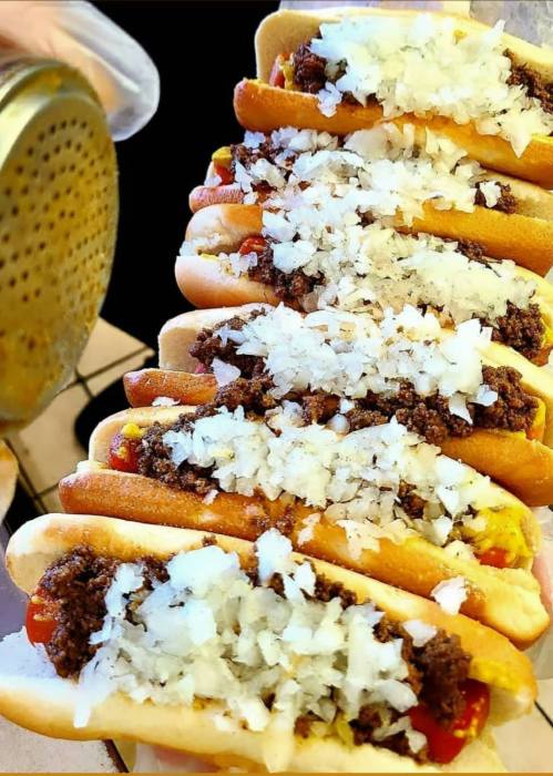 A row of hot dogs topped with chopped onions and a meat mixture, served in soft buns.