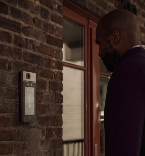 A man stands next to a modern video doorbell mounted on a brick wall, looking at the screen.