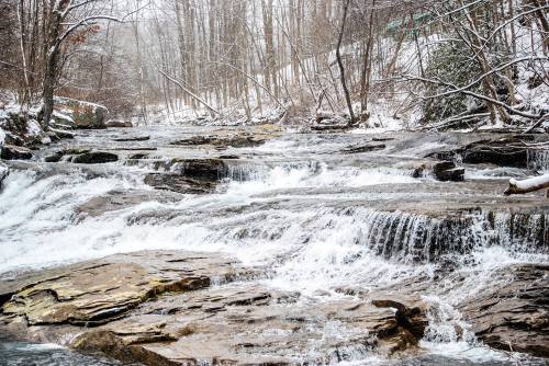 A snowy landscape featuring a flowing river over rocky terrain, surrounded by bare trees and a serene winter atmosphere.
