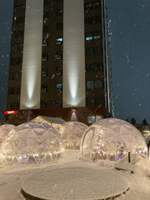 Snowy scene featuring illuminated igloo structures in front of a tall building, with snowflakes falling at night.