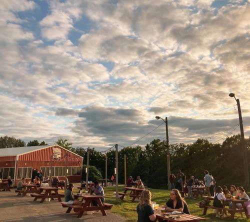Outdoor gathering at a brewery with people sitting at picnic tables under a cloudy sky. Warm evening atmosphere.