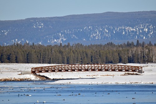 A metal bridge spans a frozen river, surrounded by snow-covered land and evergreen trees under a clear blue sky.