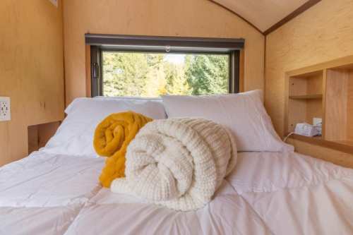 Cozy bed with white and yellow rolled blankets, set against a window with a view of trees outside.