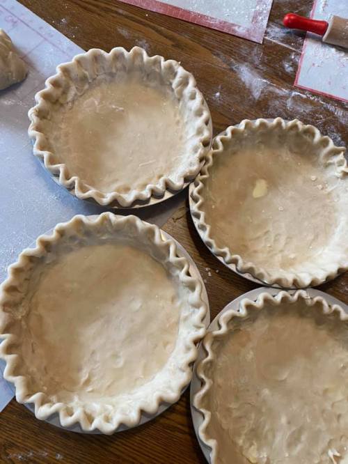 Four unbaked pie crusts with fluted edges sit on a wooden table, surrounded by flour and baking tools.
