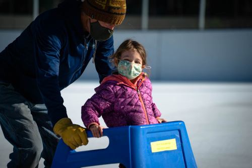 A man helps a smiling girl on ice skates using a blue skating aid, both wearing masks.