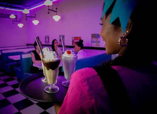 A server holds a tray of milkshakes in a retro diner with neon lights and customers in the background.