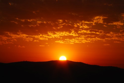 A vibrant sunset with a glowing orange sun rising above dark silhouettes of hills and a colorful sky filled with clouds.