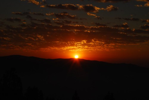 A vibrant sunset over mountains, with a golden sun peeking through clouds against a darkening sky.