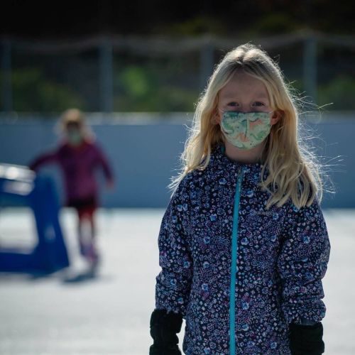 A girl with long blonde hair wearing a patterned mask and a jacket stands on an ice rink, with another skater in the background.