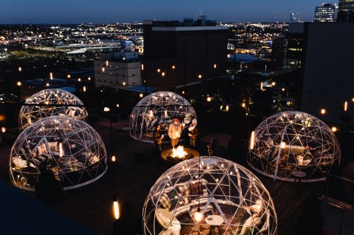 Rooftop view of illuminated igloo-like structures with people gathered around a fire, city skyline in the background at dusk.