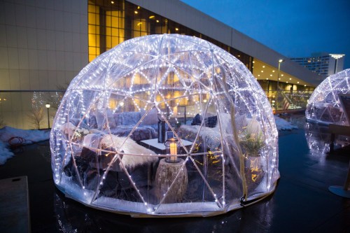 A cozy, illuminated igloo structure with soft seating, surrounded by snow and city lights at dusk.
