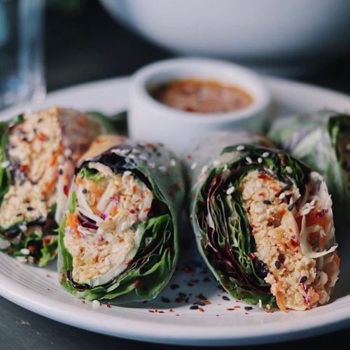 Fresh vegetable spring rolls on a plate, served with a dipping sauce and sprinkled with sesame seeds and chili flakes.