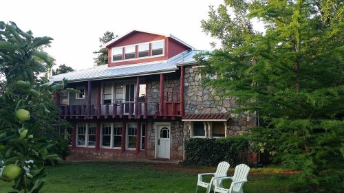 A two-story house with a stone exterior, red accents, and a metal roof, surrounded by greenery and two white chairs.