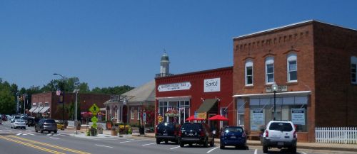 A sunny street view of a small town with brick buildings, shops, and parked cars under a clear blue sky.