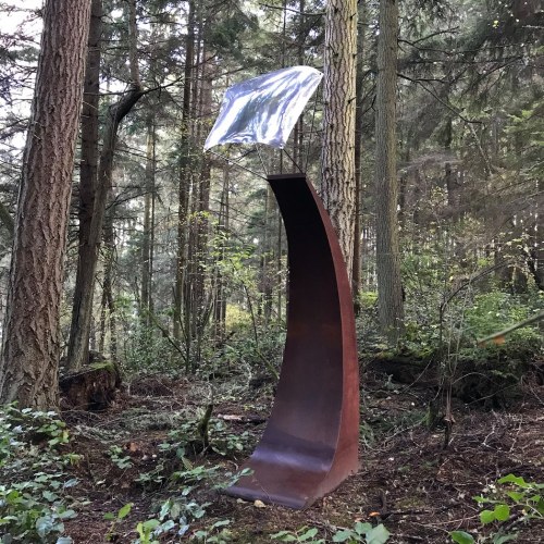 A tall, curved metal sculpture with a reflective top stands among trees in a forested area.