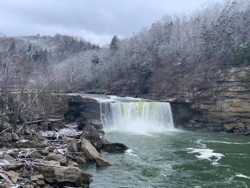 A waterfall cascades over rocky cliffs, surrounded by snow-dusted trees and a calm green river.