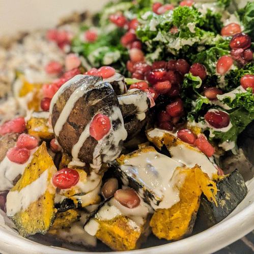 A colorful bowl featuring roasted squash, mushrooms, pomegranate seeds, and kale drizzled with a creamy dressing.