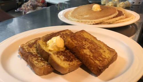 A plate of golden-brown French toast with butter, alongside a stack of pancakes in the background.
