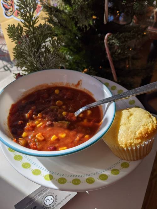 A bowl of chili with corn and beans, accompanied by a cornbread muffin, set beside a festive Christmas tree.