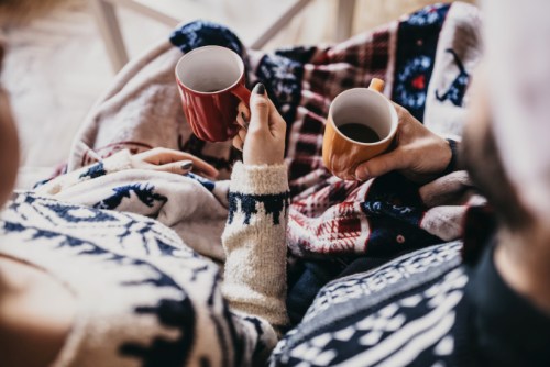 A cozy scene of two people wrapped in blankets, holding mugs of hot drinks, enjoying a warm moment together.