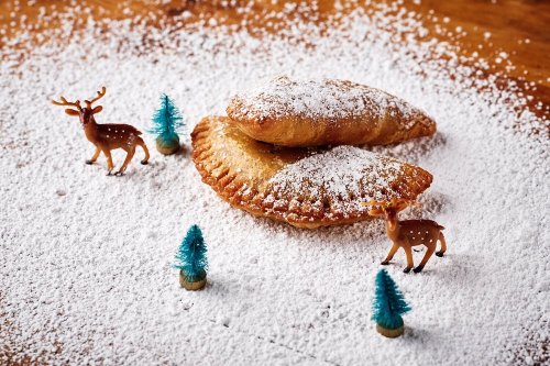 Two powdered sugar-dusted pastries surrounded by miniature trees and toy deer on a wooden surface.