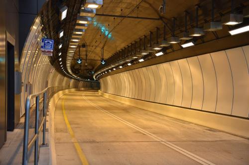 A well-lit, modern tunnel with smooth walls and a curved path, featuring directional signs and overhead lights.