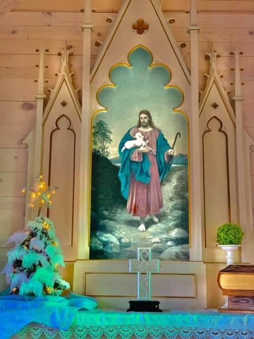 A religious painting of Jesus holding a lamb, framed by an ornate altar with a small Christmas tree and decorations.