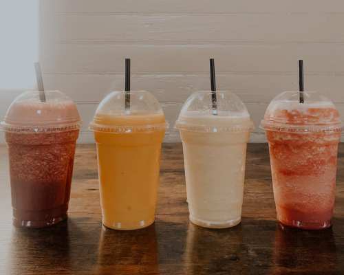 Four colorful frozen drinks in clear cups with straws, arranged on a wooden table.