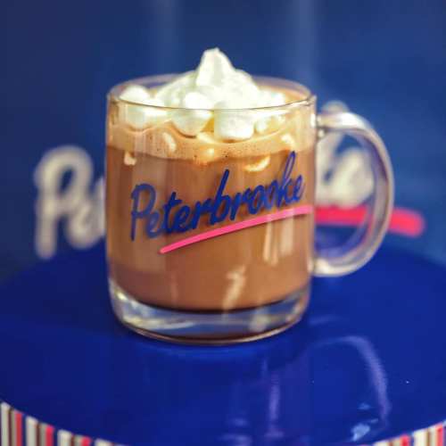 A clear mug filled with hot chocolate topped with whipped cream, featuring the logo "Peterbrooke" in blue.