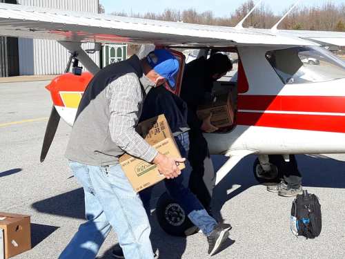 Two people loading boxes into a small airplane on a tarmac, with a hangar in the background.