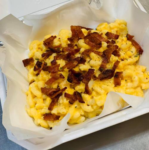 A serving of creamy macaroni and cheese topped with crispy bacon bits in a takeout container.