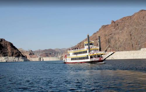 A paddleboat cruises on a river with rocky hills and a bridge in the background under a clear blue sky.