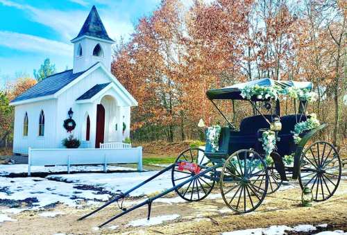 A quaint white chapel beside a horse-drawn carriage adorned with flowers, set against autumn trees and a snowy ground.