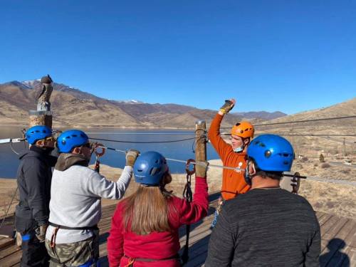 A group of people in helmets listens to an instructor on a platform with a scenic view of mountains and a lake.