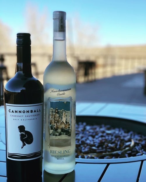 Two wine bottles, one Cabernet Sauvignon and one Riesling, sit on a table near a fire pit with a scenic background.