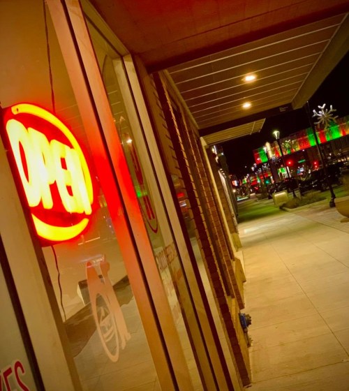 A brightly lit "OPEN" sign in a storefront window, with a festive, illuminated street scene visible outside at night.