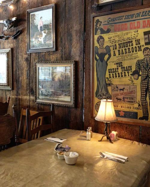 Cozy restaurant interior featuring wooden walls, vintage artwork, a table set for dining, and a lamp.