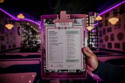 A hand holds a colorful diner menu featuring milkshakes and coffee options, with neon lights in the background.