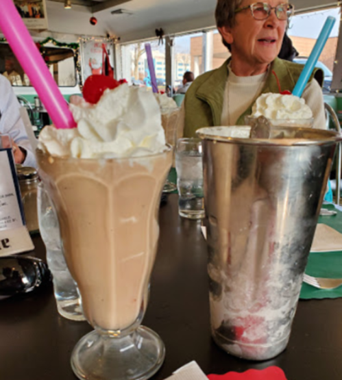 Two milkshakes topped with whipped cream and cherries, with a metal shaker and a woman in the background.