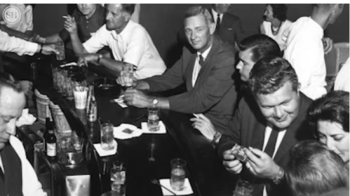 A black-and-white photo of people socializing at a bar, with drinks and snacks on the counter.