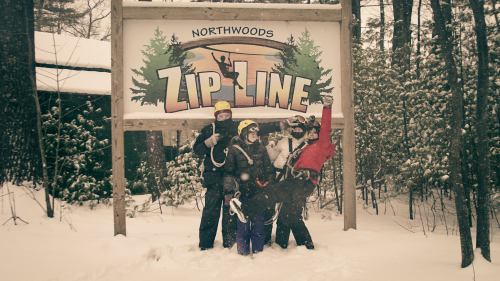 A group of four people in winter gear poses playfully in front of a "Northwoods Zip Line" sign in a snowy forest.