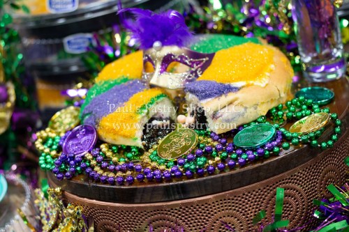 A colorful king cake decorated with purple, green, and gold, surrounded by Mardi Gras beads and coins.